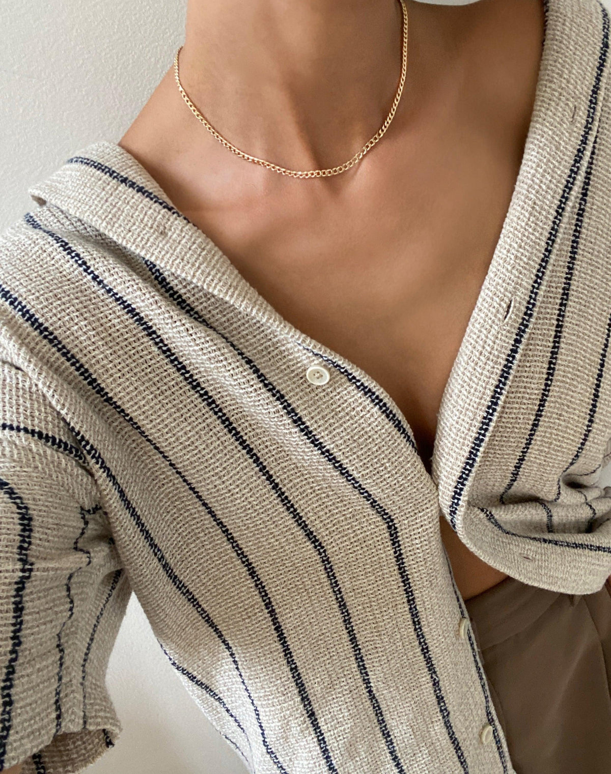 A person wears a beige, textured, button-up shirt with dark stripes over a tan top and an elegant LLUME Jewelry Marie Chain 18k gold-filled, waterproof necklace, showing it on their collarbone.