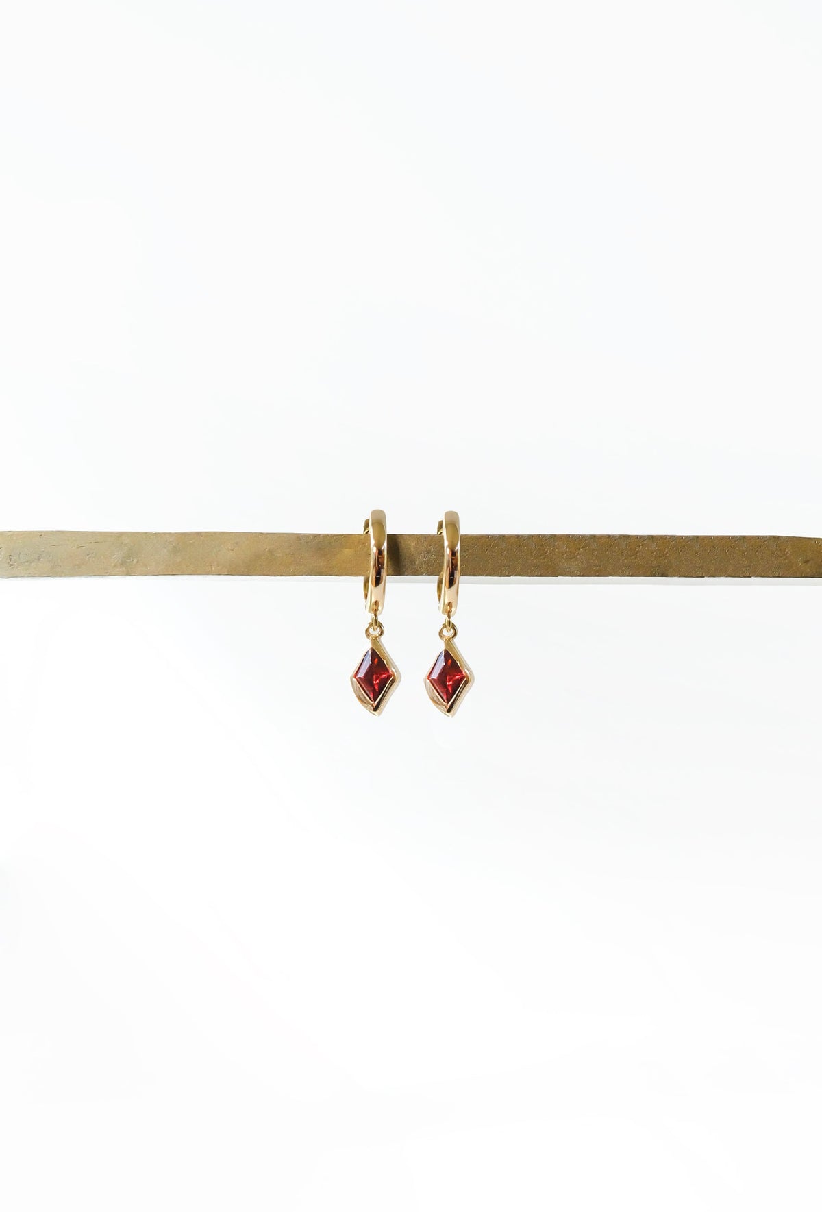 The LLUME Jewelry Birthstone Earrings feature small, square red birthstone pendants on gold-plated hooks, elegantly displayed on a horizontal brass rod against a plain white background.