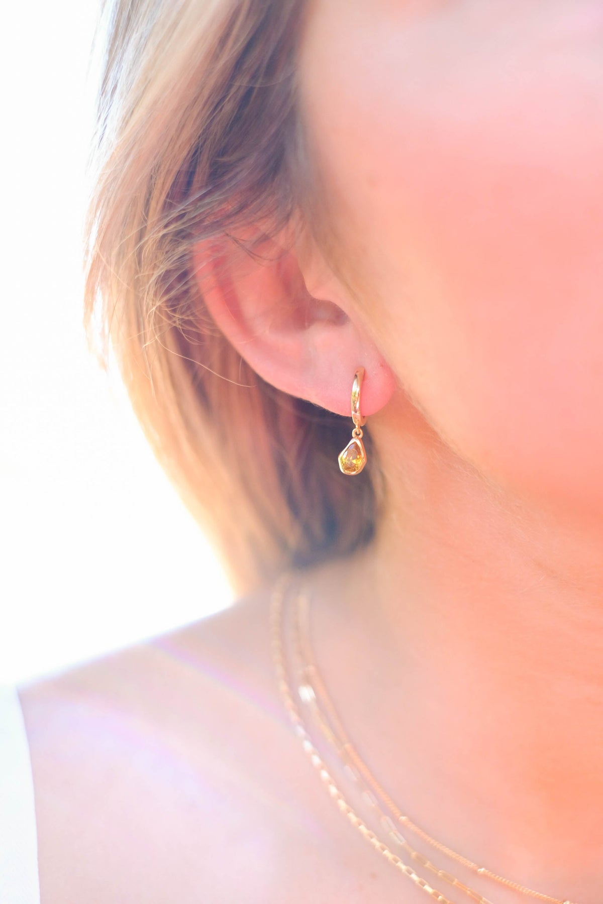 Close-up of a woman's ear wearing LLUME Jewelry's Birthstone Earrings. She also wears layered gold necklaces. The bright, softly focused background highlights the elegant gold-plated earrings.