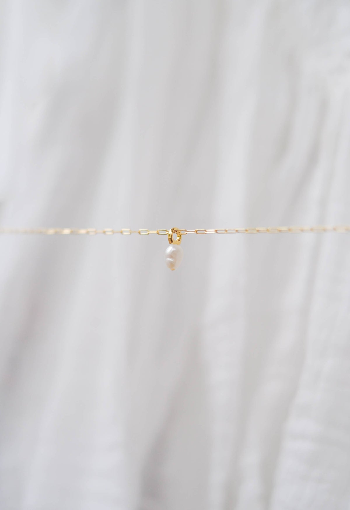 The Build a Charm Necklace by LLUME Jewelry features a delicate gold chain with a single small pearl pendant, shown on soft white fabric—ideal for layering or customizing with additional gold filled charms.