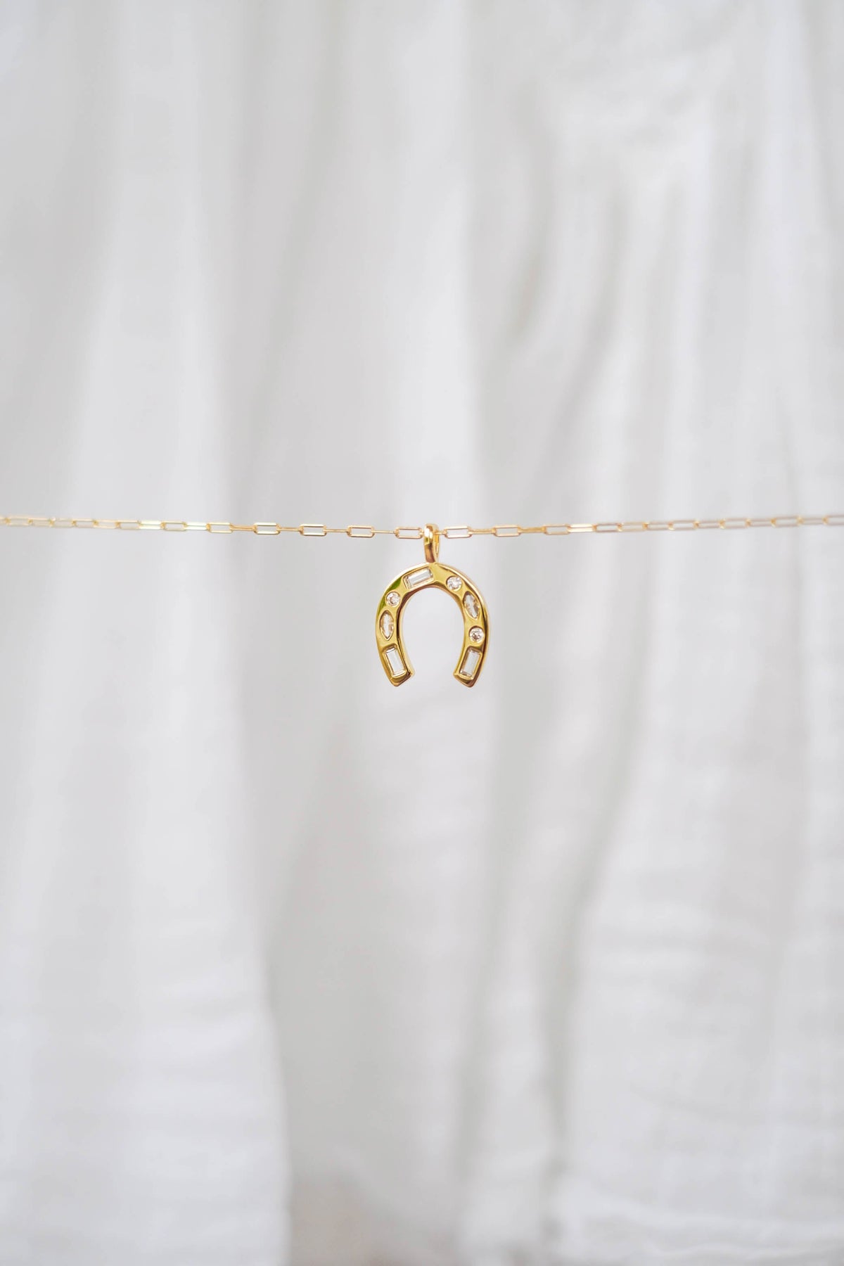 The Build a Charm Necklace by LLUME Jewelry is a delicate gold chain with a small gold horseshoe pendant adorned with tiny stones, perfect for personalizing or adding to your gold filled charm collection. Shown on a soft white fabric background.