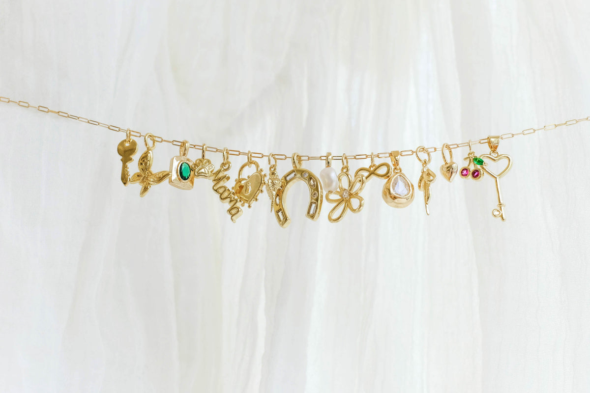 The Build a Charm Necklace by LLUME Jewelry features a gold chain adorned with gold filled charms—key, horseshoe, flower, heart, and birds—against a white fabric background.