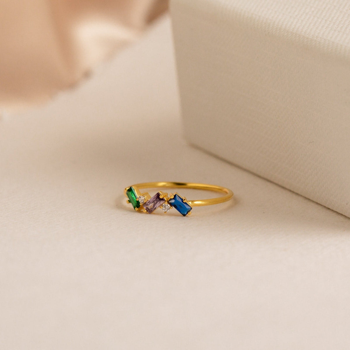Cluster Baguette Birthstone Ring in Rose Gold, featuring green, purple, clear, and blue gemstones, displayed next to a textured cream-colored box.