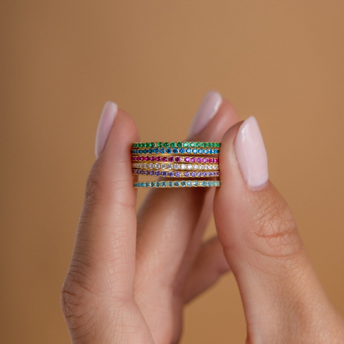 A hand holds four Eternity Birthstone Rings stacked together against a soft beige background—a customizable and elegant gemstone ring, perfect for Christmas gifting.