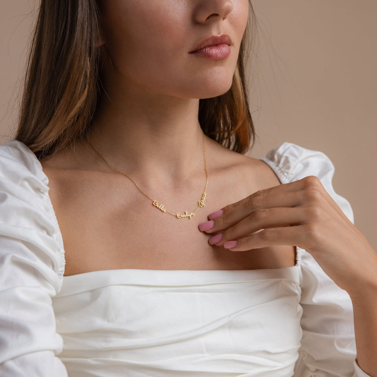 A woman in a white off-shoulder top gently touches her Dainty Tiny Multiple Name Necklace, showcasing this personalized gold accessory.