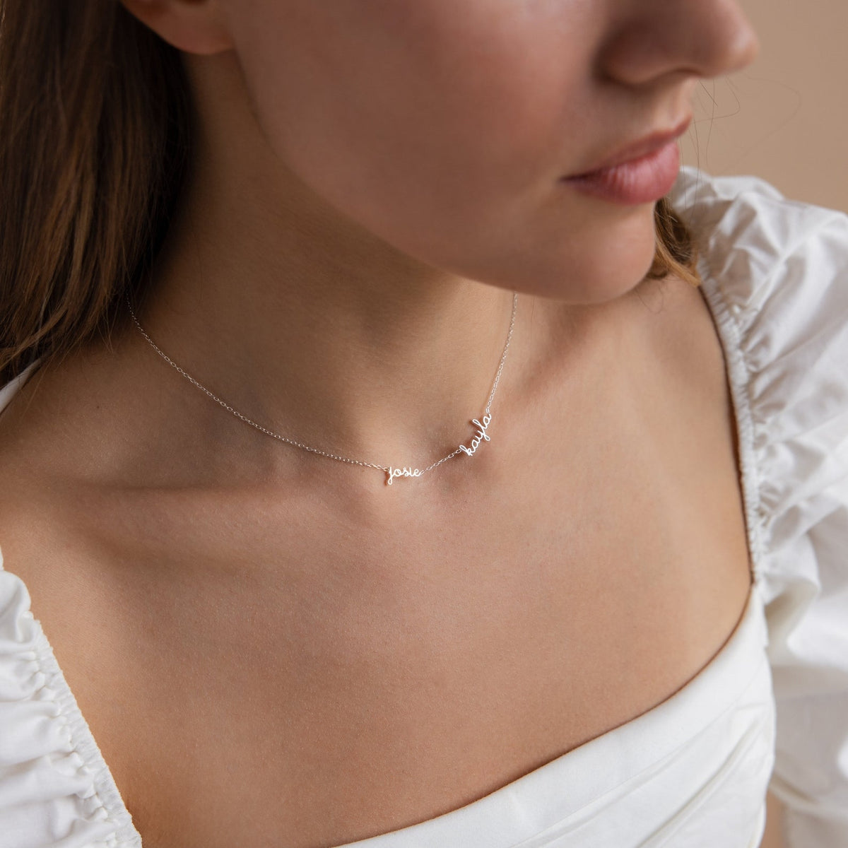 A woman wears a white off-shoulder top and a delicate necklace—a Dainty Tiny Multiple Name Necklace with personalized cursive writing, perfect for showcasing multiple names in style.
