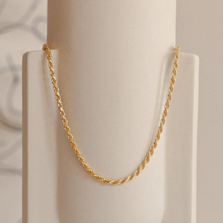 Gold rope chain necklace hanging in a graceful arc on a minimal cream ceramic vase.