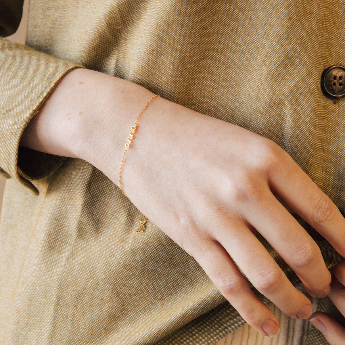 A hand wears the Petite Tiny Name Bracelet, featuring small letters, resting on a beige fabric jacket—a perfect minimal jewelry choice.