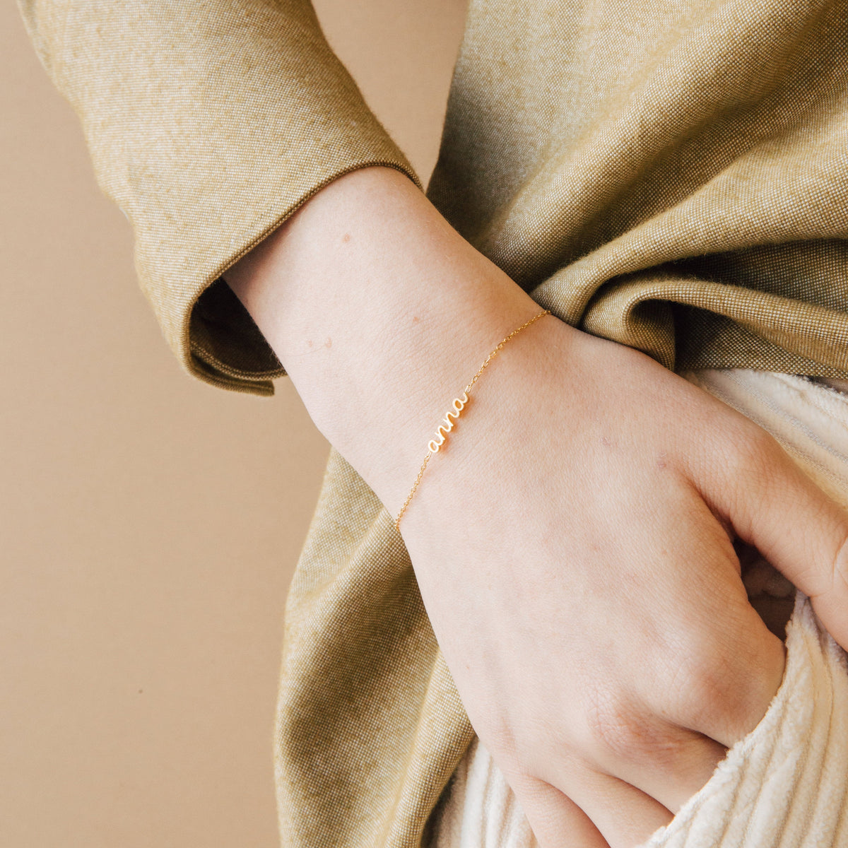 A hand wears the Petite Tiny Name Bracelet in gold, featuring the name "anna" in cursive on a delicate chain, set against a beige background.