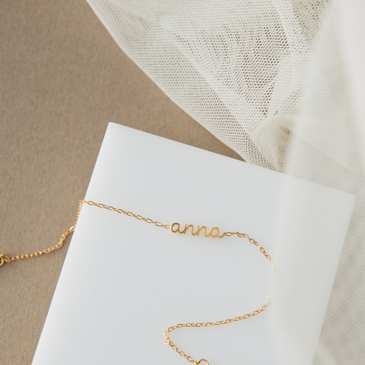 The Petite Tiny Name Bracelet showcases "anna" on a delicate chain, set against a white surface with beige fabric and mesh—ideal for minimal jewelry enthusiasts.