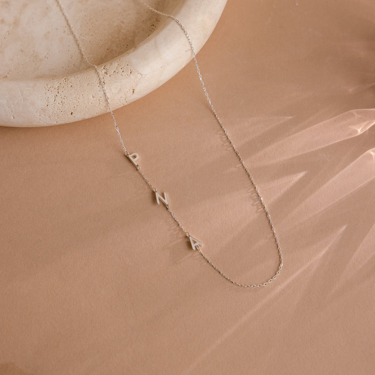 The Sideways Initial Necklace, displayed on a beige surface next to a round stone dish, features gold PNA letters—a unique and thoughtful gift for Christmas.