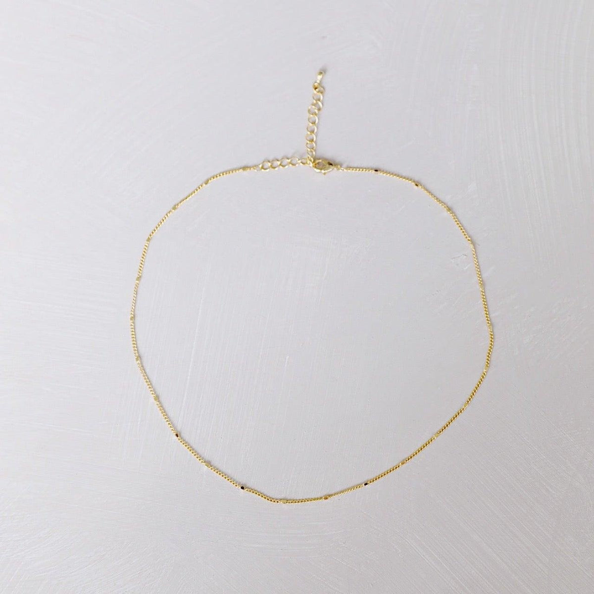 The Linden Chain by LLUME Jewelry is a delicate, dainty gold necklace with a fine, simple design, featuring an adjustable clasp and extender, crafted in 18K gold-plated finish and displayed on a light textured surface.