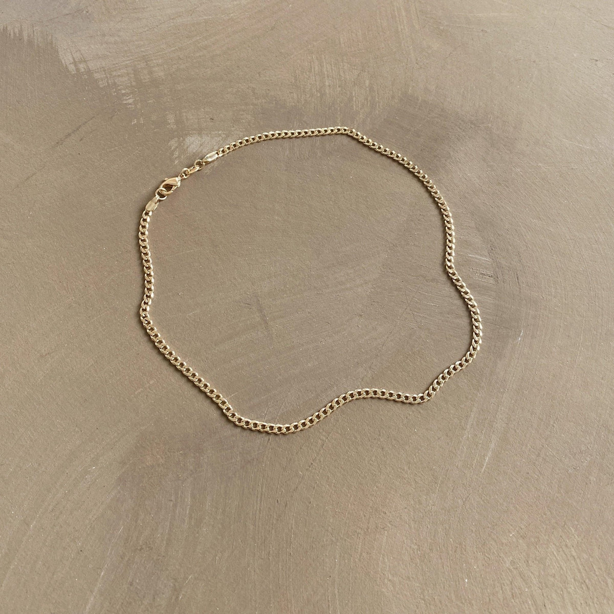 The Marie Chain by LLUME Jewelry, a thin 18k gold-filled necklace with a simple clasp, is displayed in a loose oval shape on a smooth, light brown surface.