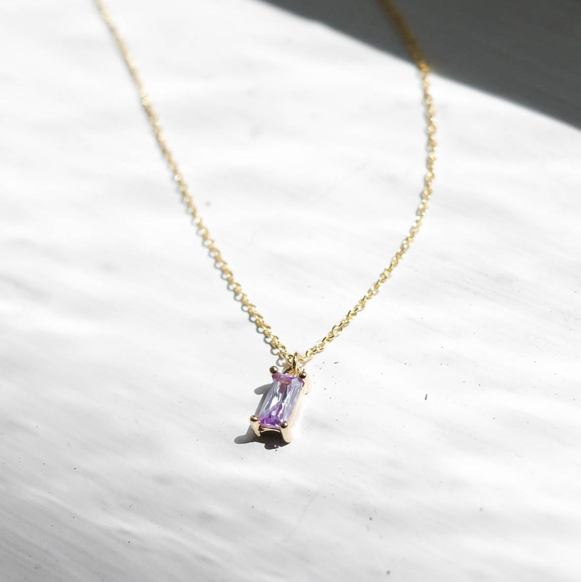 The Celia Necklace by LLUME Jewelry is a delicate gold vermeil piece featuring a small rectangular lilac gemstone pendant, displayed on a white textured surface in natural light.