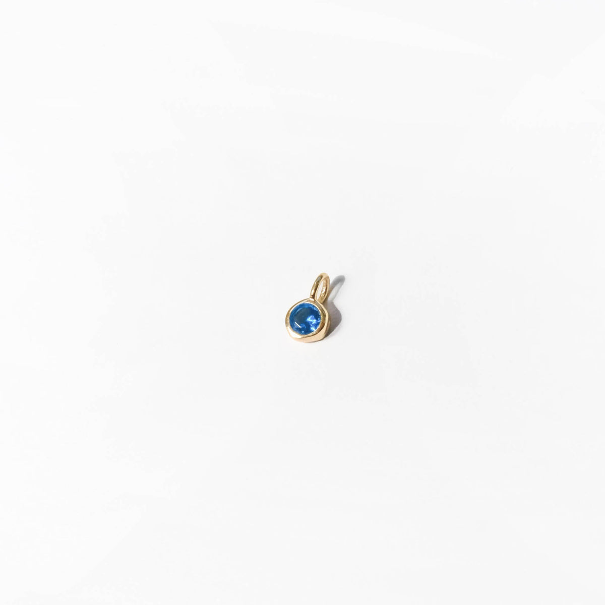 The Build a Birthstone Necklace by LLUME Jewelry features a small gold pendant with a round blue gemstone at the center, elegantly displayed against a plain white background.