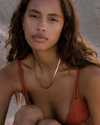 Close-up portrait of a model wearing a gold herringbone necklace and small gold hoop earrings in a terracotta bikini top, outdoors