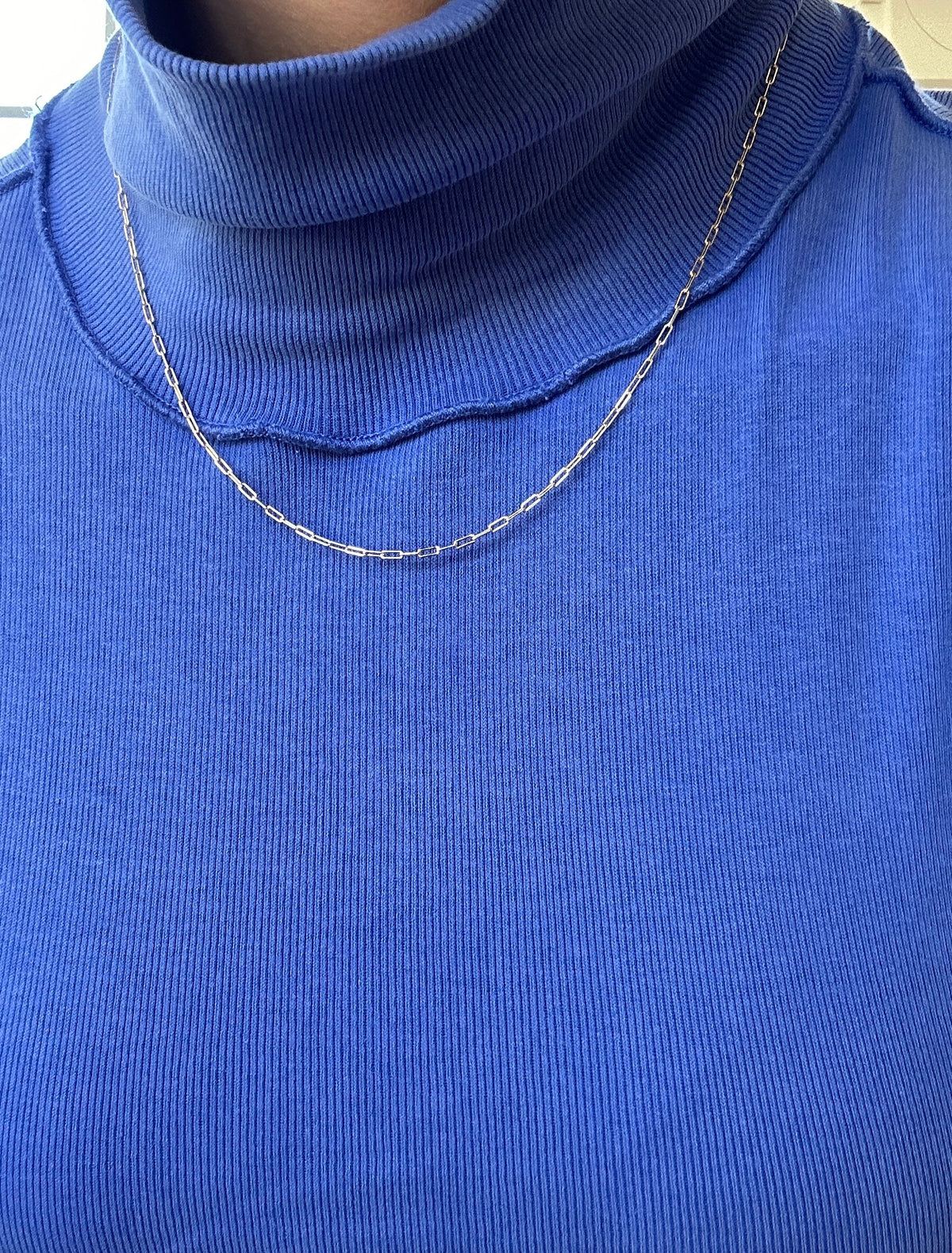 Someone wears a bright blue ribbed turtleneck top paired with the Adelene Paperclip Chain by LLUME Jewelry, a thin and delicate gold-plated necklace. The photo highlights the chest and neckline.