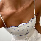 A person wears a white, floral-patterned camisole and high-waisted white pants, accessorized with LLUME Jewelry's Golden Hour Chain necklace. The cropped photo highlights the upper torso.