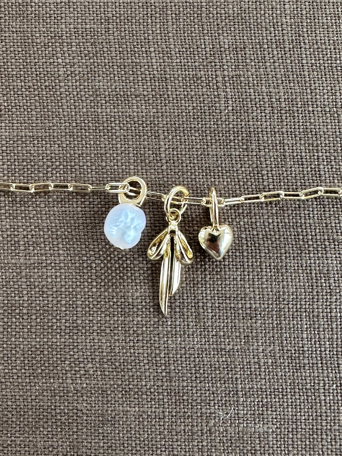 A gold chain bracelet with three gold-filled charms—a translucent flower, a wing, and a heart—from LLUME Jewelry rests on textured brown fabric, inspiring you to create your own Build a Charm Necklace or pair it with your favorite piece.