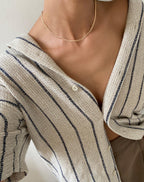 A person wears a beige, textured, button-up shirt with dark stripes over a tan top and an elegant LLUME Jewelry Marie Chain 18k gold-filled, waterproof necklace, showing it on their collarbone.
