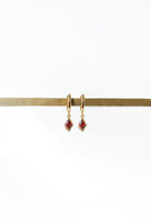 The LLUME Jewelry Birthstone Earrings feature small, square red birthstone pendants on gold-plated hooks, elegantly displayed on a horizontal brass rod against a plain white background.