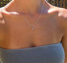 A person outdoors in sunlight wears a gray strapless top and the LLUME Jewelry Build a Birthstone Necklace—a thin silver chain with an oval pink pendant—around their neck.