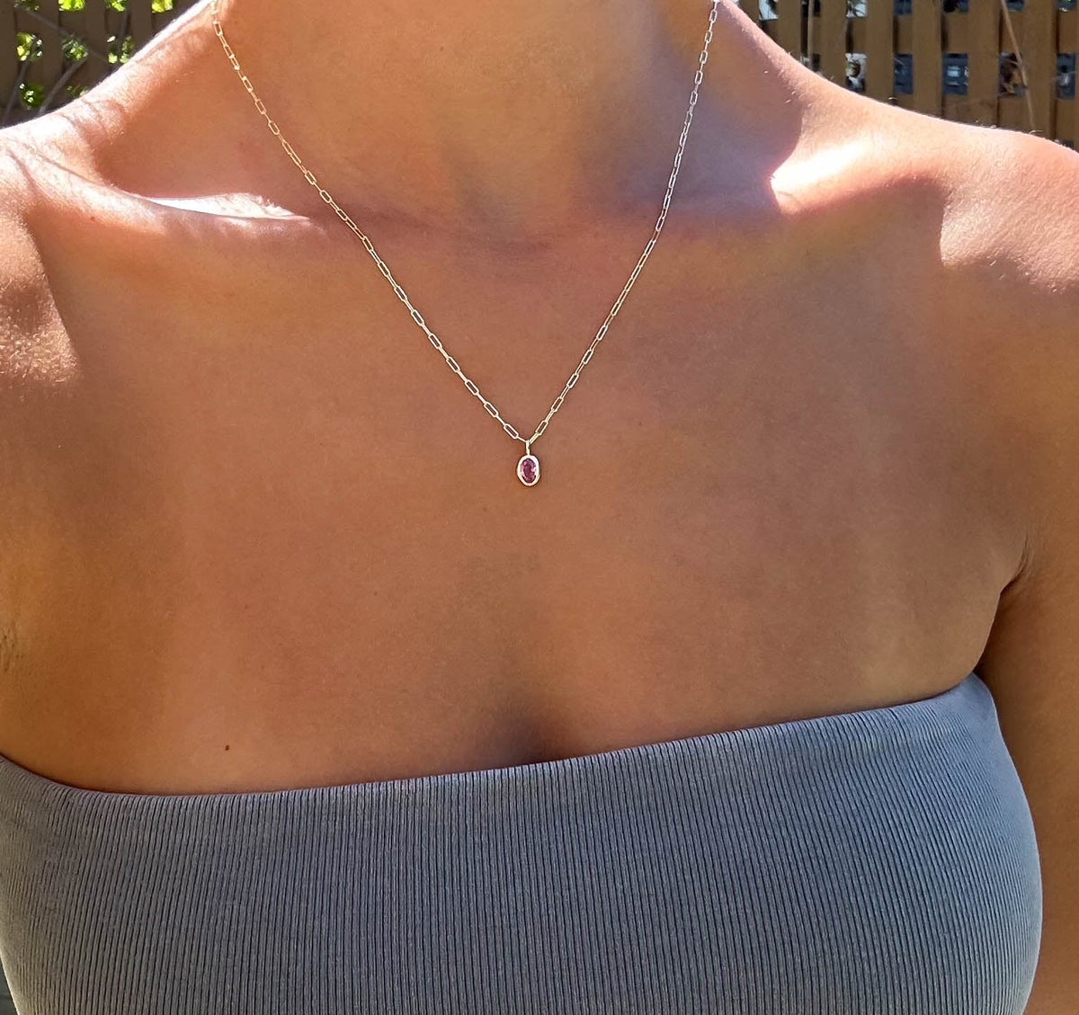 A person outdoors in sunlight wears a gray strapless top and the LLUME Jewelry Build a Birthstone Necklace—a thin silver chain with an oval pink pendant—around their neck.