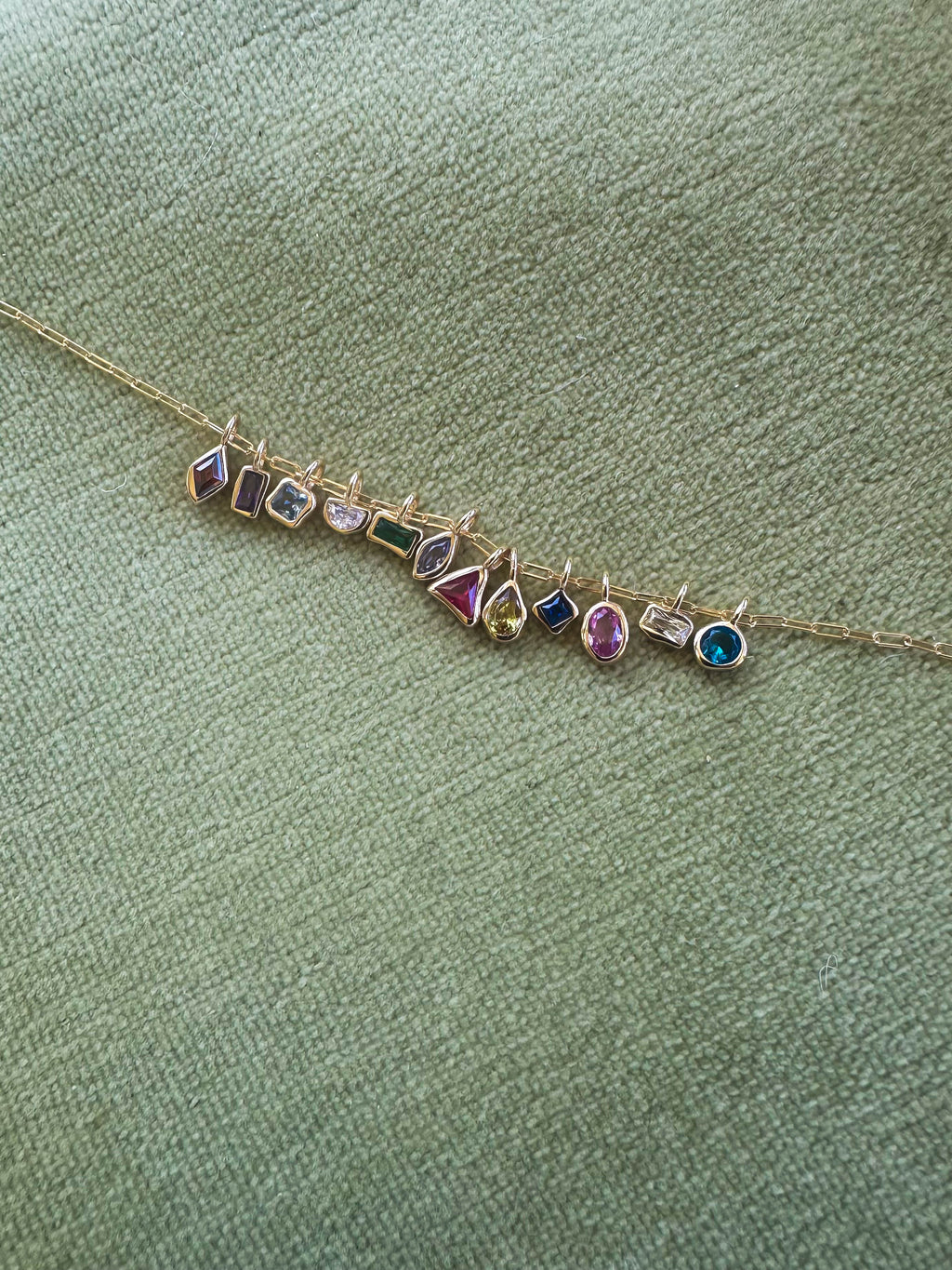 The LLUME Jewelry Build a Birthstone Necklace features gold plated charms and colorful gemstones in unique shapes, set against a textured light green fabric background.