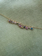 The LLUME Jewelry Build a Birthstone Necklace features gold plated charms and colorful gemstones in unique shapes, set against a textured light green fabric background.