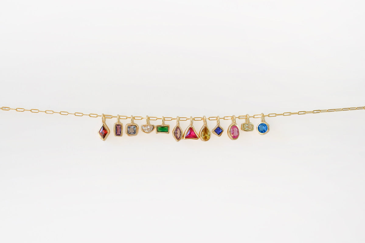 The Build a Birthstone Necklace by LLUME Jewelry features a gold chain with eleven unique, gold-plated gemstone charms in different shapes and settings, showcased against a plain white background.