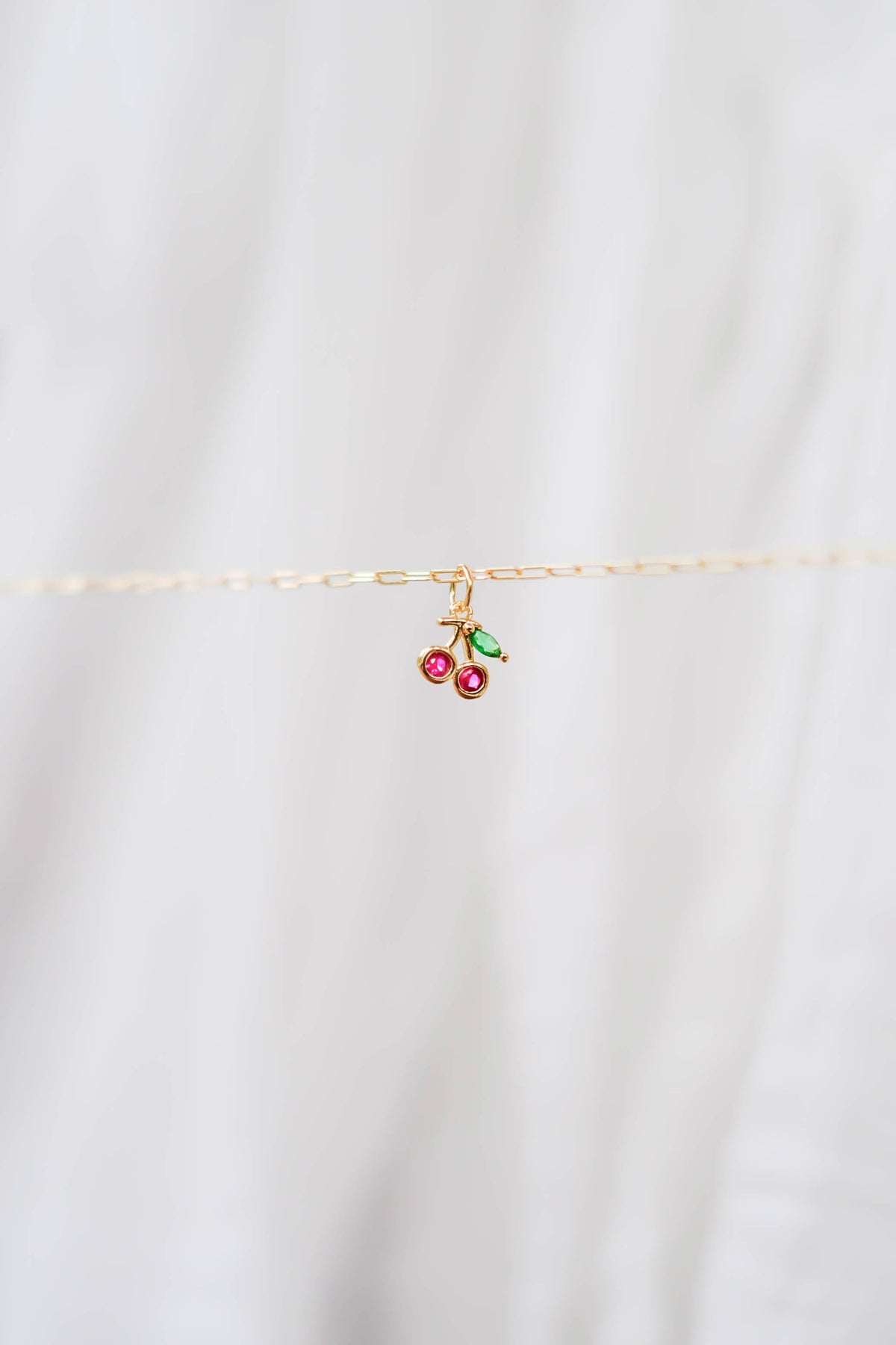 The LLUME Jewelry Build a Charm Necklace features a delicate gold chain with a cherry pendant of two red beads and green leaves, perfect for personalizing your necklace collection. Shown on a soft white background.