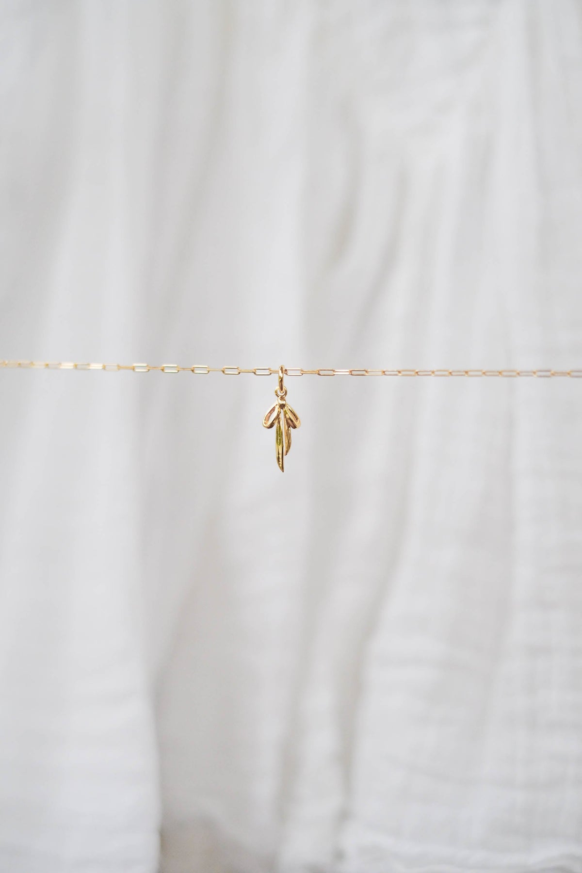 The Build a Charm Necklace by LLUME Jewelry features a delicate gold chain with a small gold leaf pendant, displayed on soft white fabric—ideal for layering or adding to your gold filled charm collection.