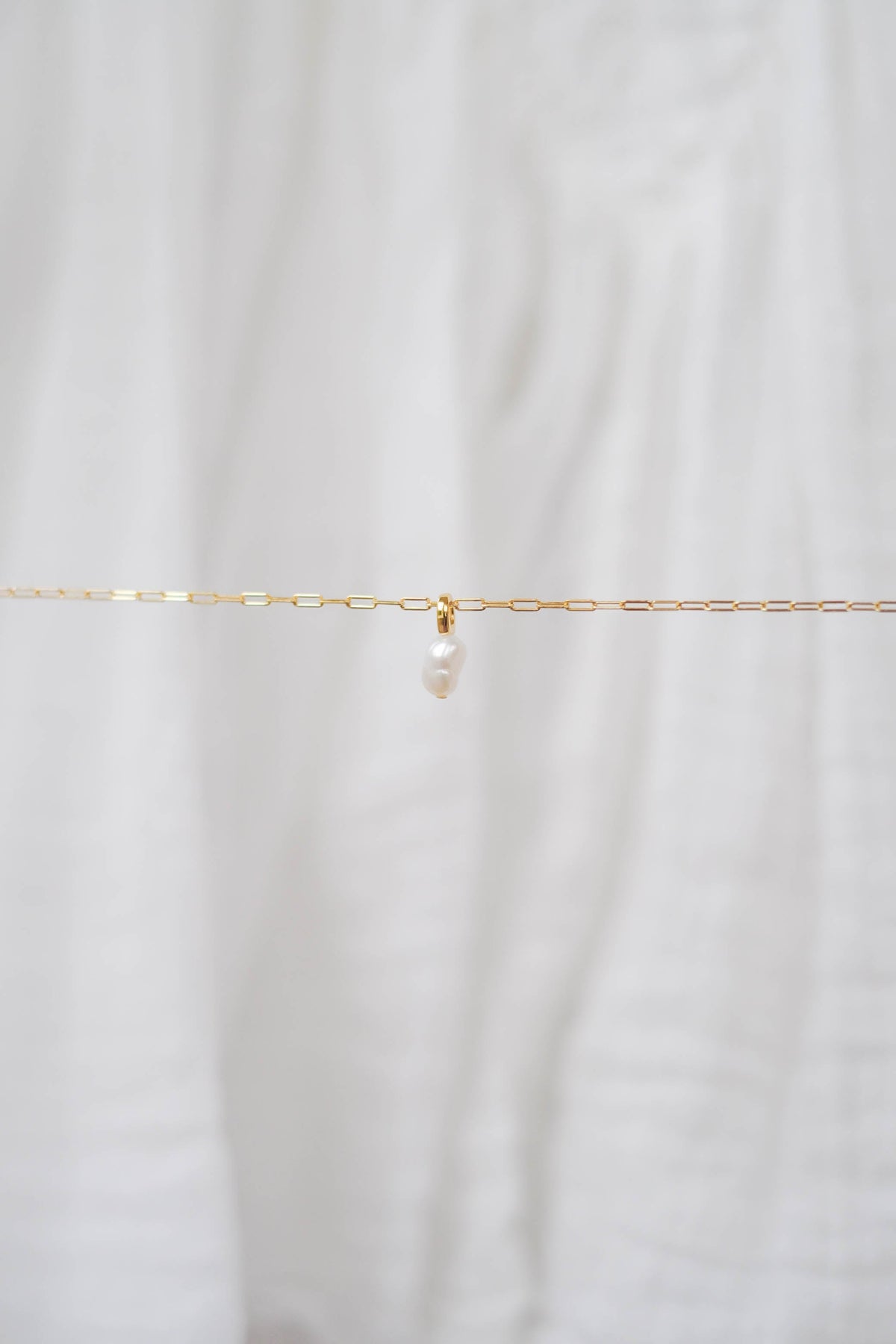 The Build a Charm Necklace by LLUME Jewelry features a delicate gold-filled chain with a single white pearl pendant—ideal for layering or adding your favorite charms. Displayed elegantly against soft white fabric.