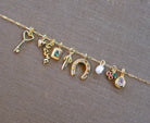 A gold chain by LLUME Jewelry showcases assorted gold filled charms—heart key, “mama” script, blue stone, and more—on a brown fabric backdrop, inspiring you to personalize your own Build a Charm Necklace.