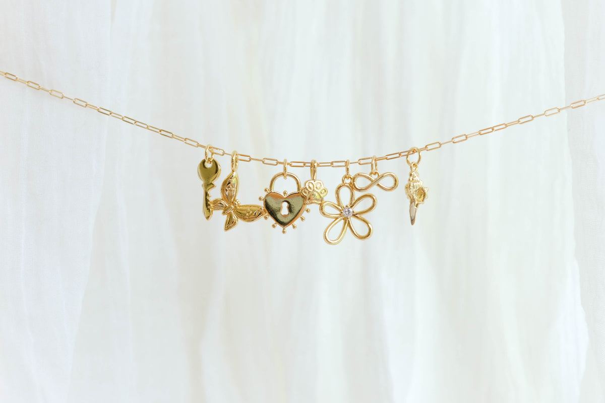 The Build a Charm Necklace by LLUME Jewelry features six unique gold filled charms—key, leaves, heart, paw print, gemmed flower, and ornate pendant—on a gold chain, elegantly displayed on soft white fabric for a truly personalized accessory.
