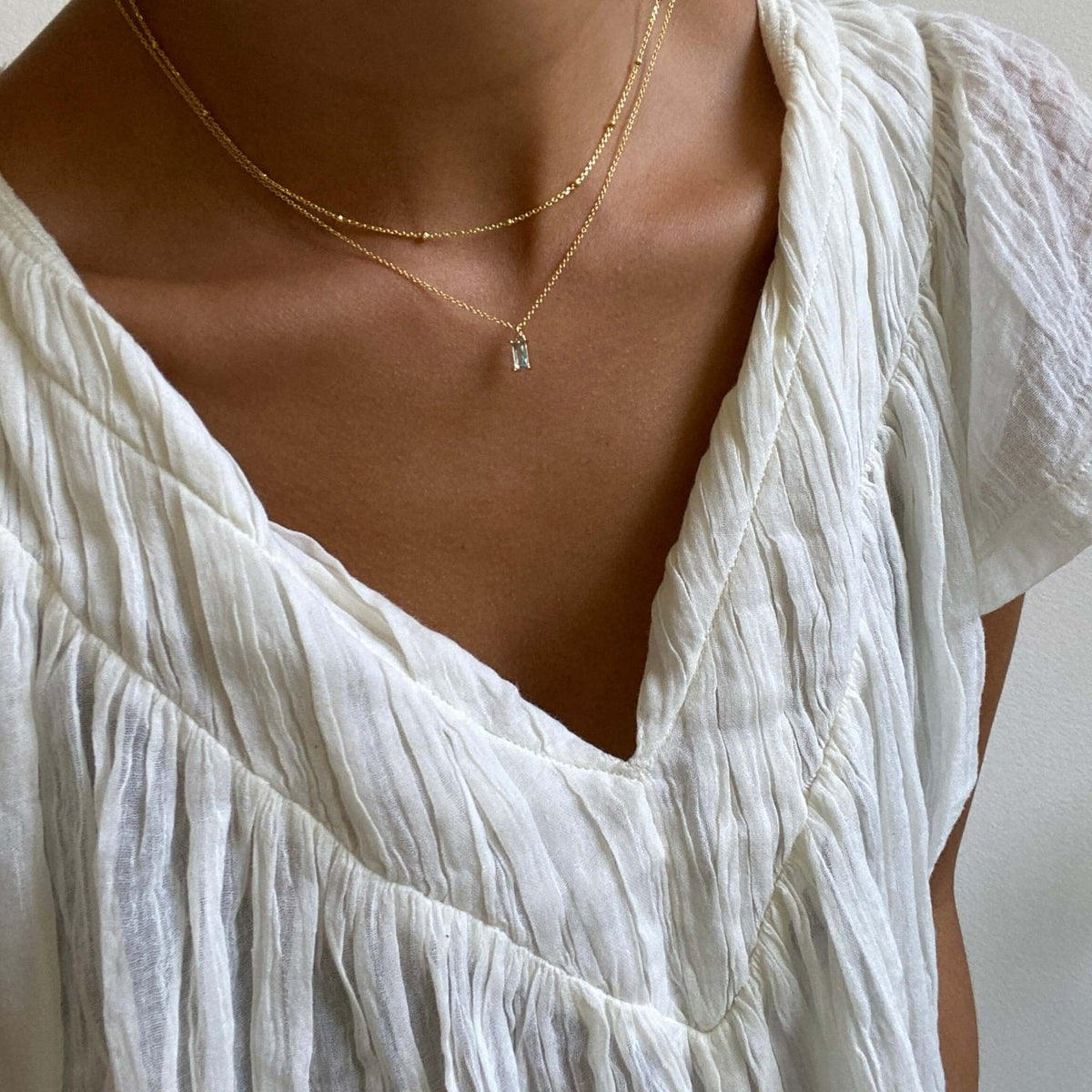 A person wears a white, textured v-neck blouse with two delicate necklaces—a gold-plated LLUME Jewelry Daylight Necklace and a tiny charm necklace—against a neutral background.