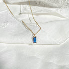 The Taylor Necklace by LLUME Jewelry, featuring a rectangular sapphire blue gemstone pendant with gold plating, is displayed on white textured fabric.