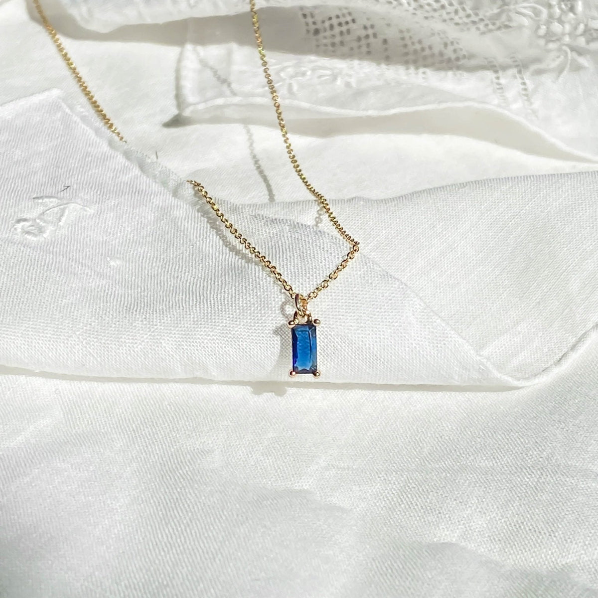 The Taylor Necklace by LLUME Jewelry, featuring a rectangular sapphire blue gemstone pendant with gold plating, is displayed on white textured fabric.