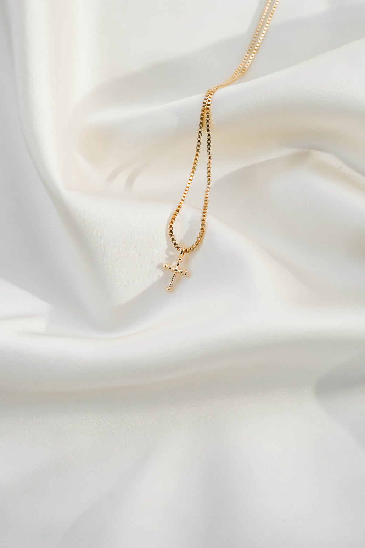 The LLUME Jewelry Cross Necklace, featuring a gold plated chain, is beautifully displayed on soft white silky fabric.