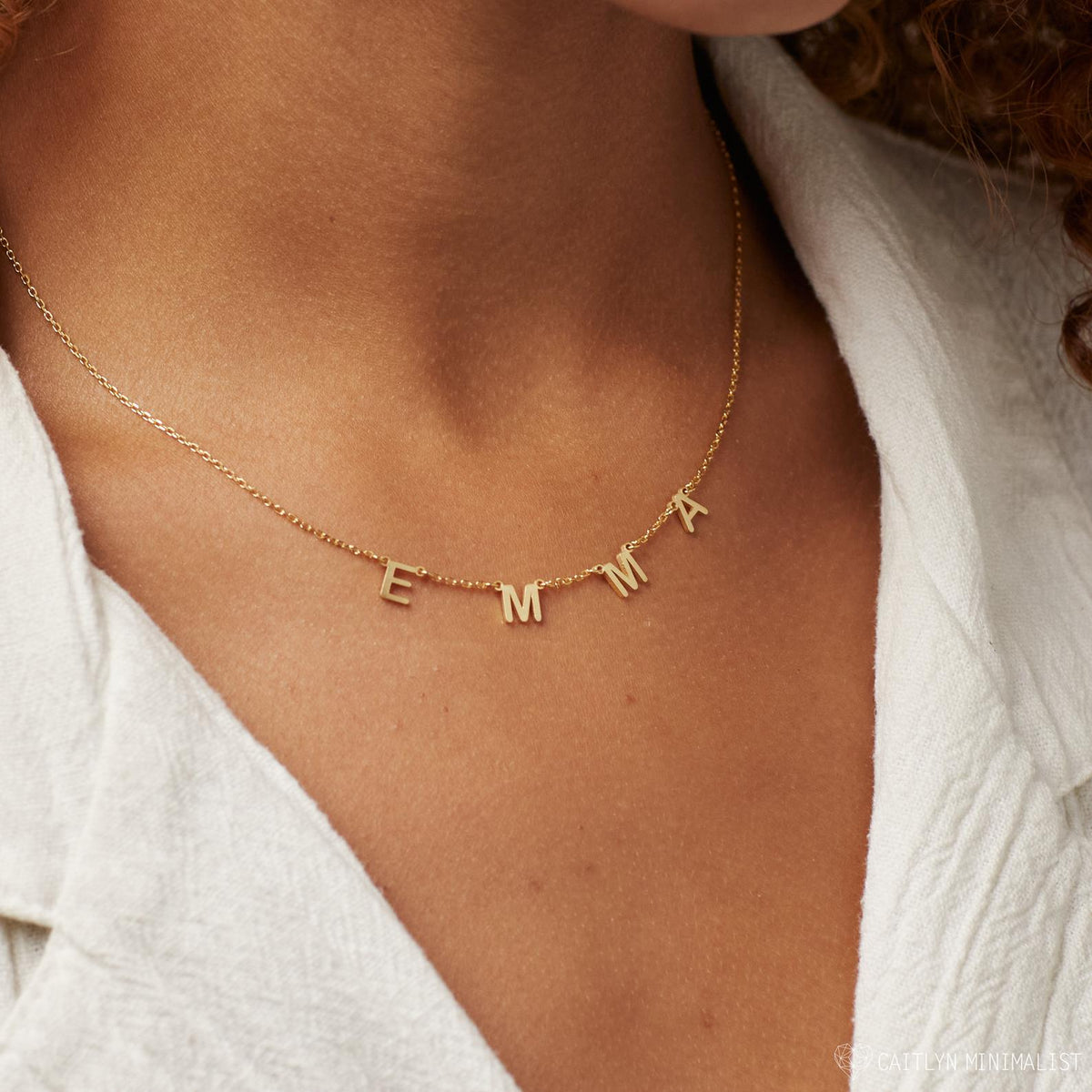 The Alice Letter Name Necklace in Rose Gold spells EMMA and is worn with a white textured shirt—making it a stylish personalized jewelry piece or thoughtful Christmas gift.