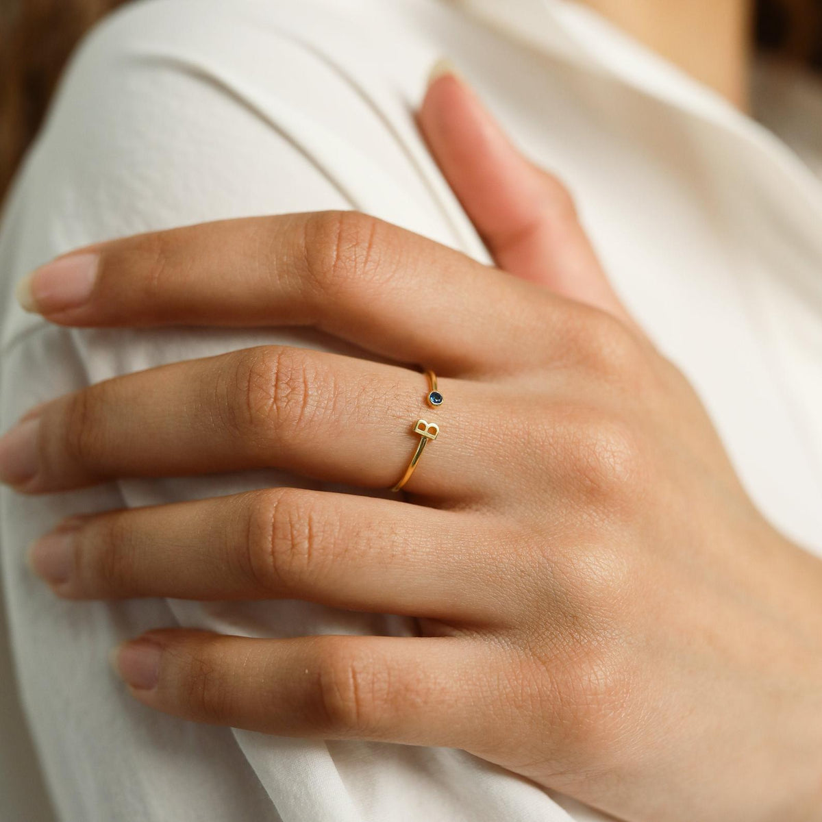 A hand with the Modern Initial Birthstone Ring, featuring a delicate blue gem and gold finish, rests on a white shirt—an elegant, customizable ring ideal as a Christmas gift.