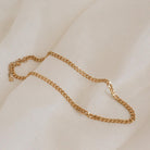 A close-up shot of a chunky gold curb chain necklace with flat, interlocking links, draped gently across a soft ivory-colored fabric