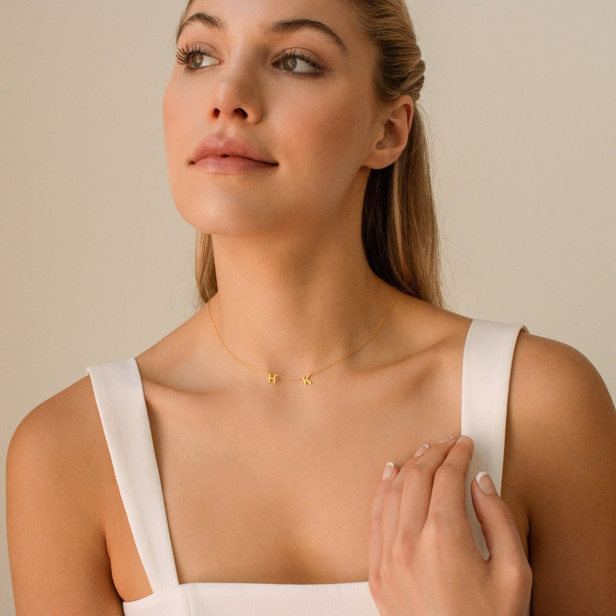 A woman in a white dress wears the Alice Dainty Initial Necklace with H and K initials, touching her collarbone—an ideal personalized jewelry piece for a thoughtful Christmas gift.