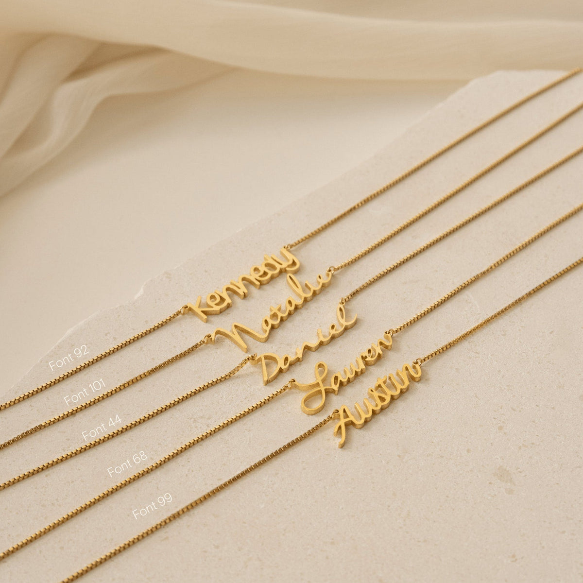 Five Callie Box Chain Name Necklaces in 18K Gold are displayed in various fonts on a light beige surface, with soft fabric in the background.