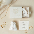 A beige surface displays a jewelry box with gold rings, two small pouches, and the Tiny Summer Name Necklace in a delicate font, accompanied by a note that says "Enjoy!".