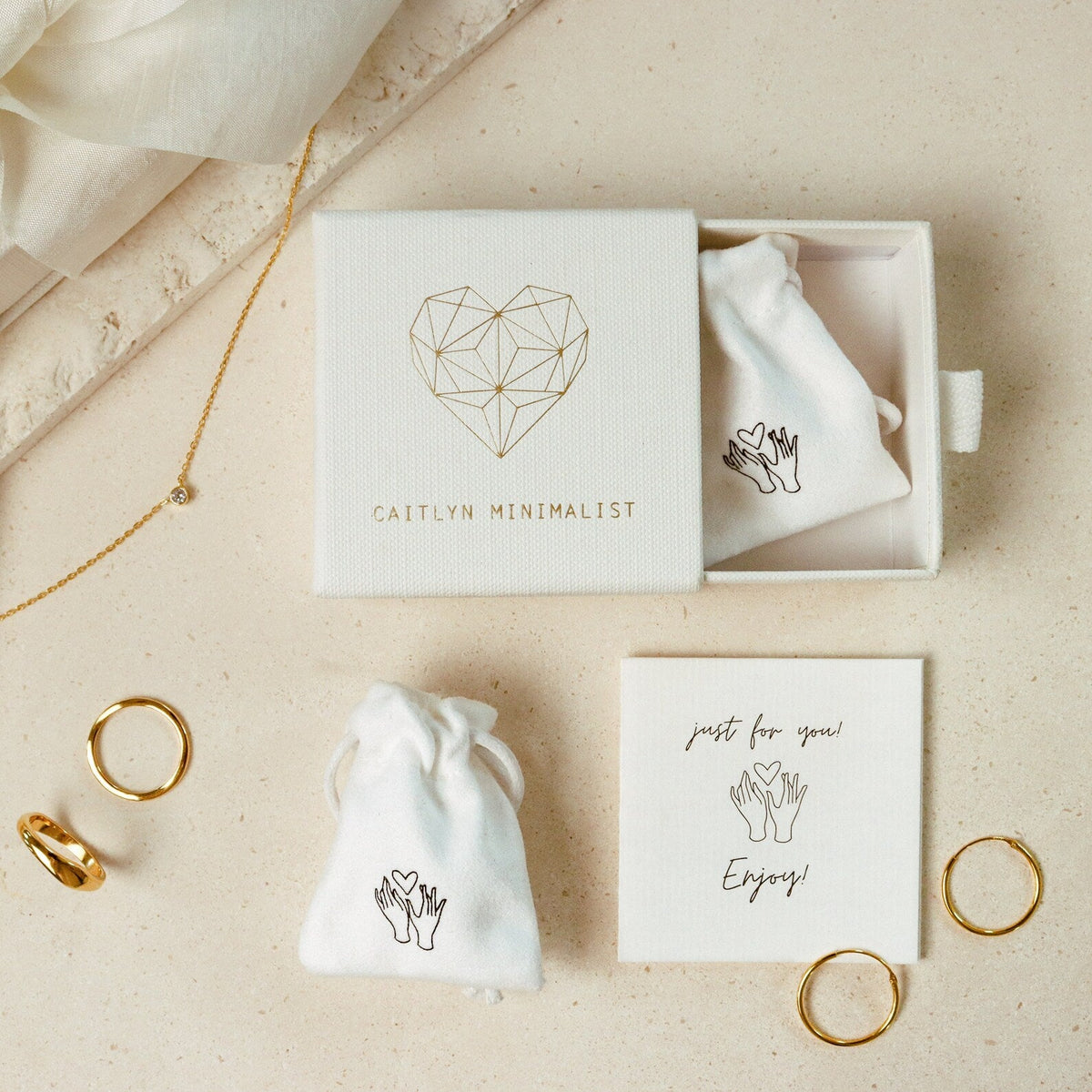 A beige surface displays a jewelry box with gold rings, two small pouches, and the Tiny Summer Name Necklace in a delicate font, accompanied by a note that says "Enjoy!".
