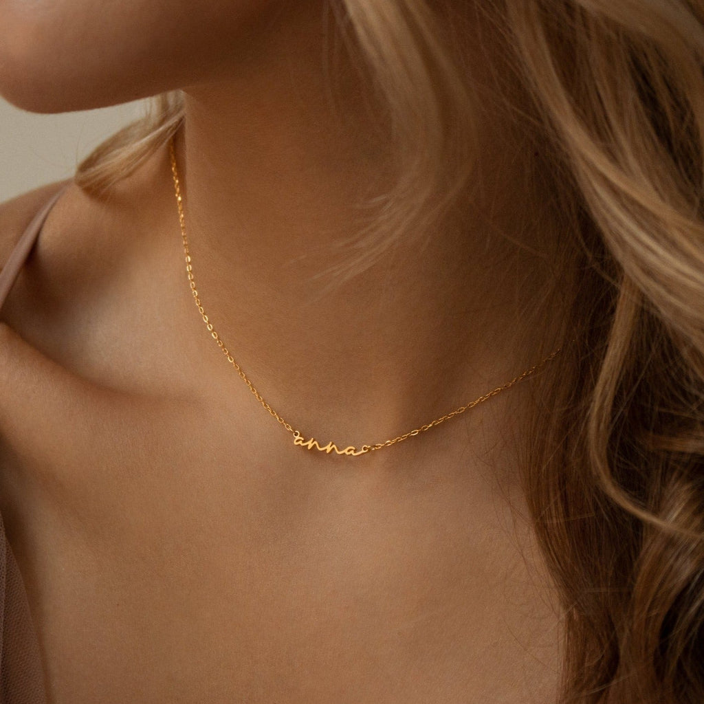 A woman with wavy blonde hair wears the Tiny Summer Name Necklace, featuring her custom name in delicate gold cursive.