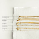 Flat-lay image showing ten labeled gold chains side-by-side on a white cloth, each labeled by style 