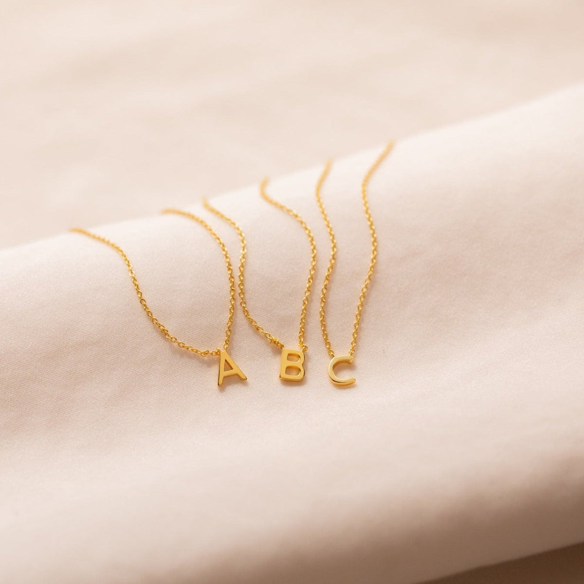 Three Dainty Initial Necklaces with gold letter pendants A, B, and C displayed on soft cream fabric—perfect as a customizable accessory or thoughtful Christmas gift.