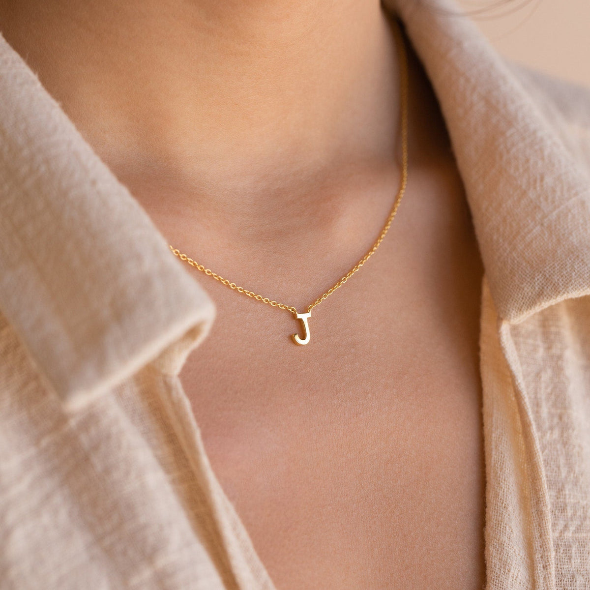 The Dainty Initial Necklace features a gold letter J pendant, making it a perfect Christmas gift, shown worn over a beige textured shirt.
